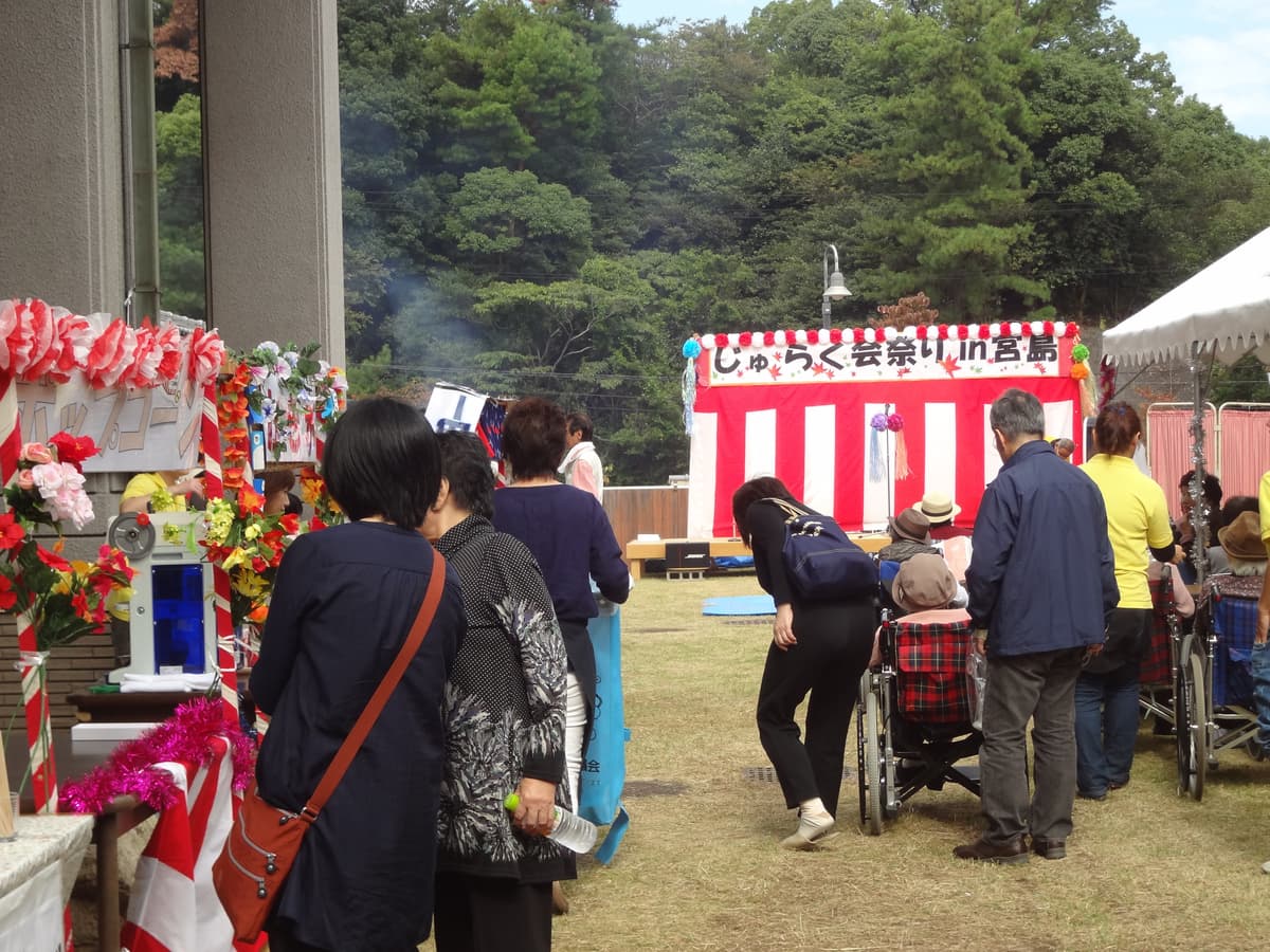 施設の夏祭りイベント。入居者と来園者が屋外で盆踊りや露店を楽しんでいる様子。